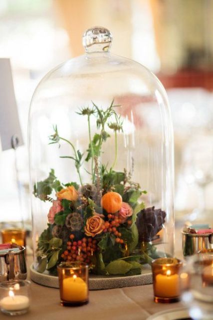 a catchy wedding centerpiece of a cloche with berries, bright blooms, an artichoke and greenery for summer or fall