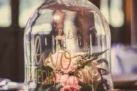 a catchy wedding centerpiece of a cloche with gold calligraphy, neutral blooms and greenery and a tree slice