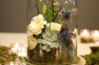 a catchy wedding centerpiece of a cloche with moss, white blooms, greenery, thistles and succulents