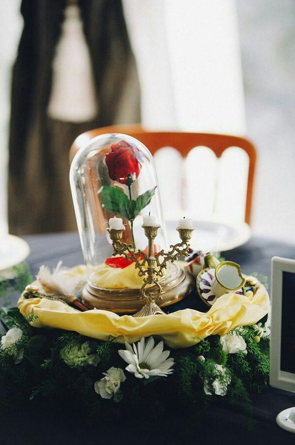 a chic Beauty and the Beast wedding centerpiece with greenery, whiet blooms, gold fabric, candles and a single red rose in a cloche