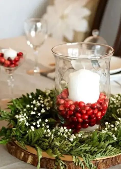 a chic rustic winter wedding centerpiece of a wood slice, greenery, baby’s breath and a glass with cranberries and a candle