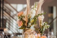 a cluster wedding centerpiece of vases with neutral and peachy blooms and grass, a couple of cloches with lights