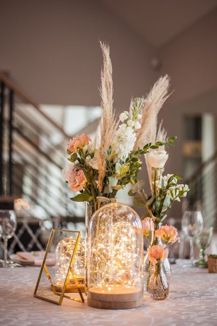 a cluster wedding centerpiece of vases with neutral and peachy blooms and grass, a couple of cloches with lights