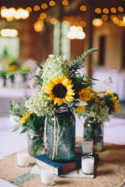 a cool rustic wedding centerpiece of jars with greenery, green hydrangeas and sunflowers plus candles around is an easy solution
