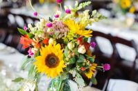 a cute and colorful rustic wedding centerpiece of a jar with sunflowers, red, pink blooms, greenery and berries plus baby’s breath around is cool