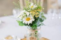 a cute rustic wedding centerpiece of a wood slice, a jar with daisies and some wildflowers plus eucalyptus and a wooden heart