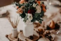 a fall wedding centerpiece of tree slices, a pumpkin with a number, pinecones and nuts, a floral arrangement in a white jar