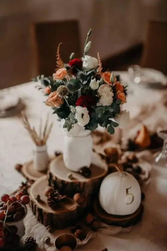 a fall wedding centerpiece of tree slices, a pumpkin with a number, pinecones and nuts, a floral arrangement in a white jar