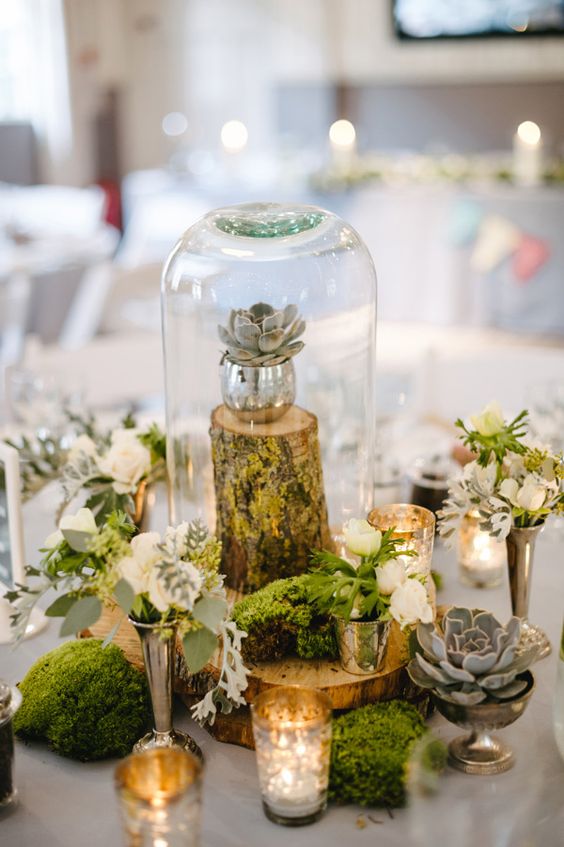 a lovely wedding centerpiece of moss, a cloche with a succulent on a stand, white blooms and silver glasses