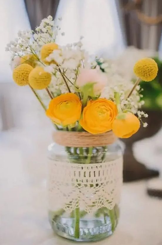 a mason jar wrapped with a doily with a floral arrangement with yellow ranunculus, baby's breath and billy balls