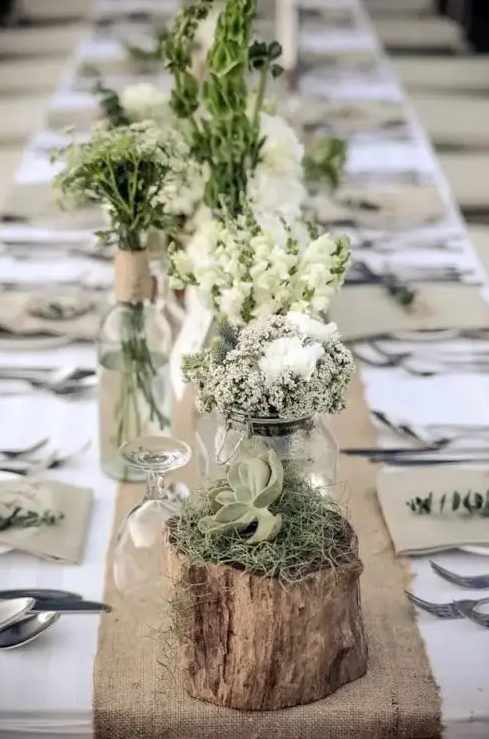 a neutral barn wedding tablescape with a burlap table runner, jars with white blooms and greenery and logs with moss and succulents