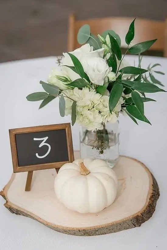 a pretty and elegant rustic fall wedding centerpiece of a wood slice, a pumpkin, white roses and hydrangeas and greenery and a chalkboard number