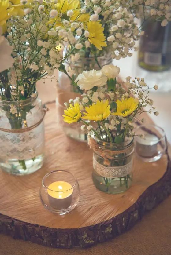 a relaxed rustic wedding centerpiece of a wood slice, candles, jars wrapped with lace, baby's breath, yellow daisies and white carnations