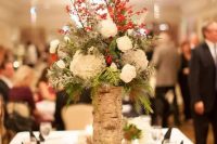 a rustic Christmas wedding centerpiece of white blooms, thistles, berries, twigs and greenery and a vase wrapped with bark