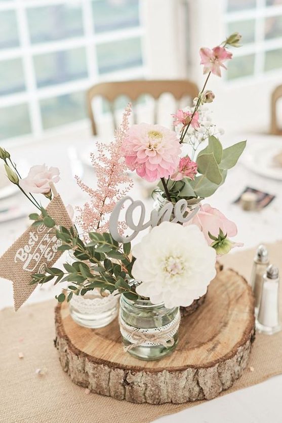 a rustic cluster wedding centerpiece of a wood slice, jars with pink and white blooms, greenery and pink astilbe, a table number and a burlap mark