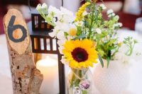 a rustic cluster wedding centerpiece with sunflowers, white blooms, a candle lantern and a tree branch with a table number