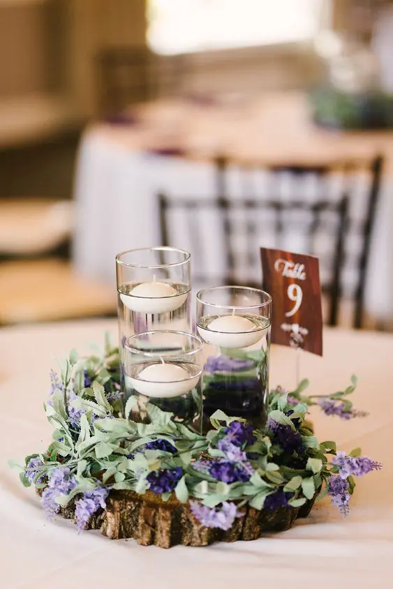 a rustic summer wedding centerpiece of a tree slice with greenery and purple blooms and floating candles plus a table number