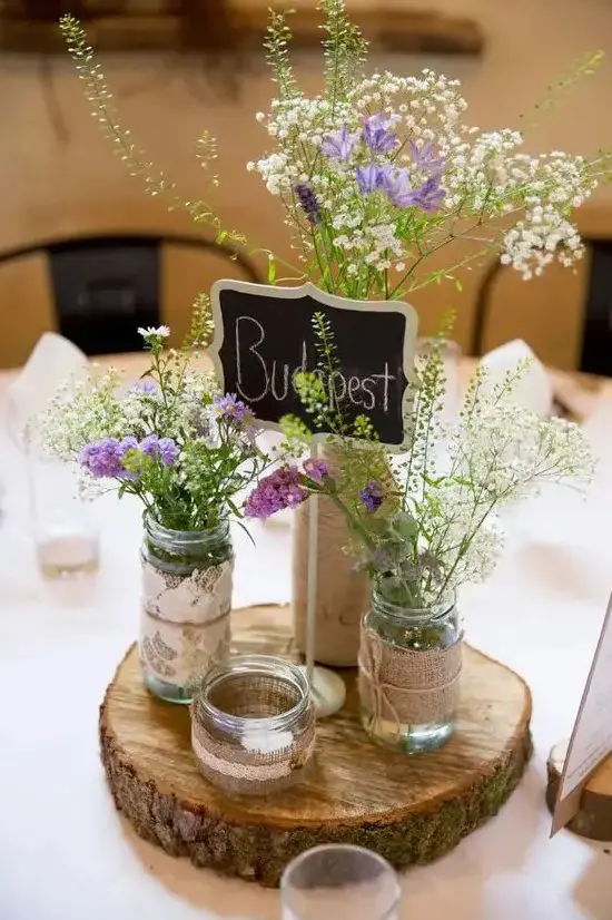 a rustic wedding centerpiece of a tree slice, mason jars with greenery, white and purple wildflowers, a chalkboard table name