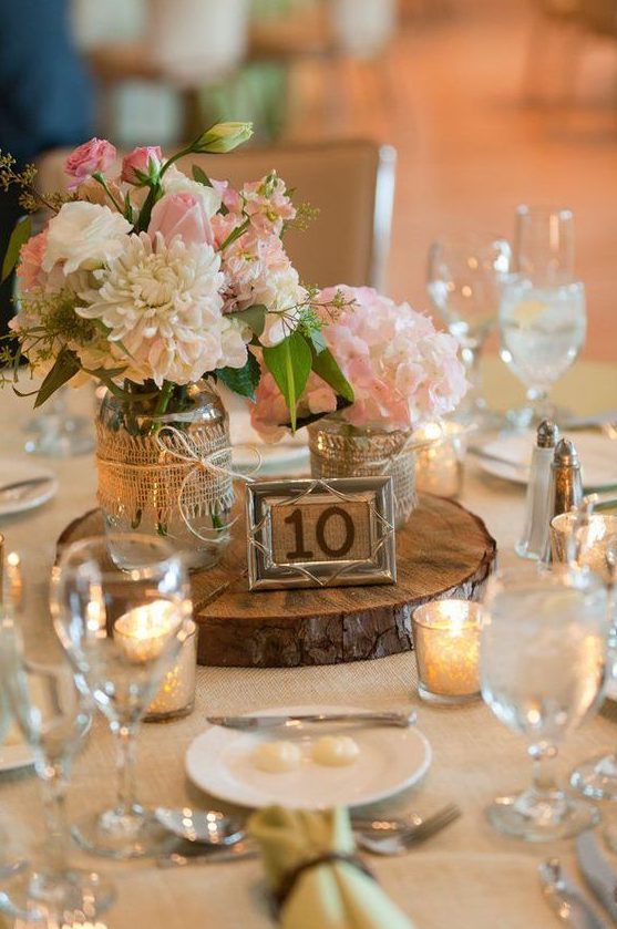 a rustic wedding centerpiece of a wood slice, a table number, pink and neutral blooms in jars wrapped with burlap