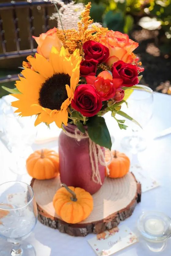 a simple and colorful wedding centerpiece of a red jar with sunflowers, red roses, dried grasses and pumpkins ona wood slice