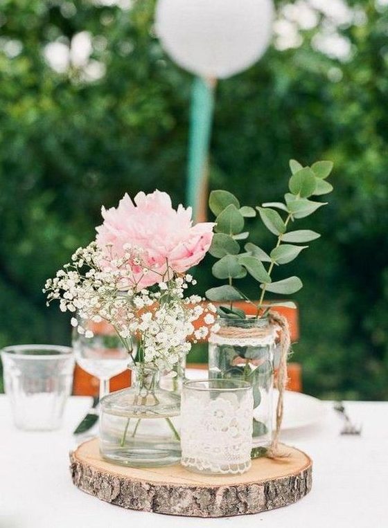 a simple rustic wedding centerpiece of a wood slice, a candleholder in lace, jars and bottles with greenery, a peony and baby's breath