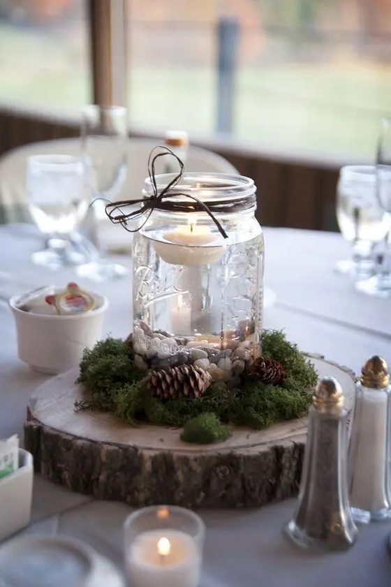 a simple wedding centerpiece of a wood slice, moss, pinecones and a jar with pebbles and a floating candle is an amazing rustic idea