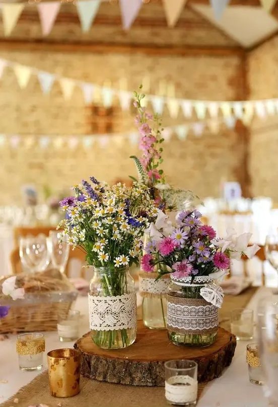 a summer barn wedding centerpiece of a wood slice, jars wrapped with burlap and lace and bright wildflowers plus candles
