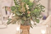 a textural rustic centerpiece of various foliage and some white blooms, the jar is wrapped with twine and a tag
