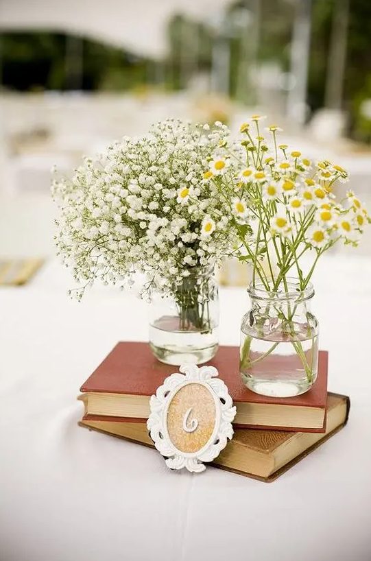 a vintage rustic wedding centerpiece of a stack of books and two vases, one with baby's breath and the second with daisies