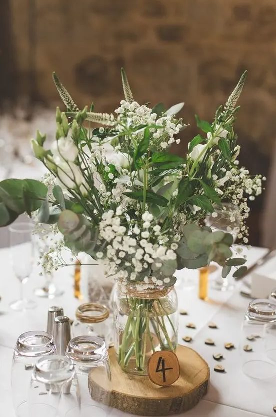 an airy barn wedding centerpiece of a wood slice, a wood slice table number and neutral blooms and greenery in a jar
