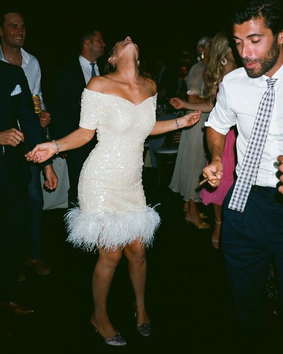 a white sequin off the shoulder over the knee dress with feathers is ideal for a wedding reception for a fun glam wedding