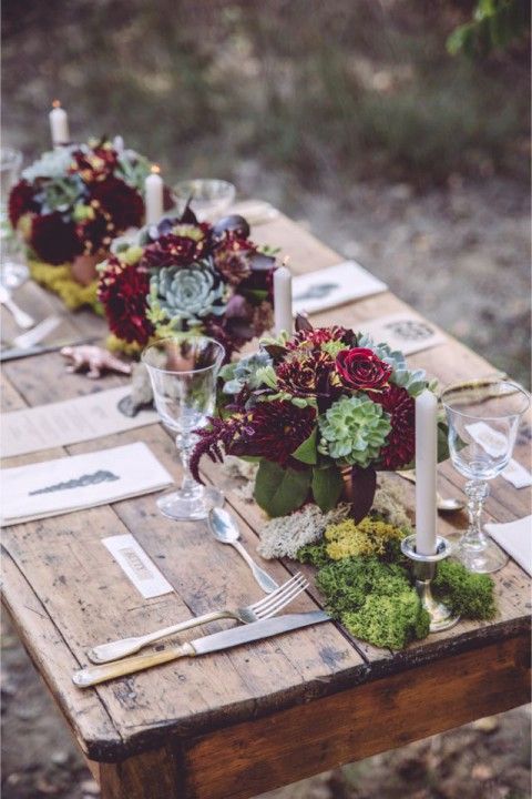 a wooden table with moss and bold burgundy and green centerpieces, candles and simple cutlery