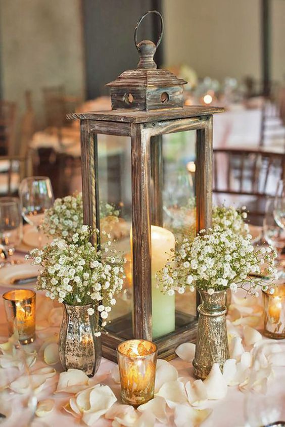 a wooden candle lantern, baby's breath arrangements in mercury glass vases and flower petals