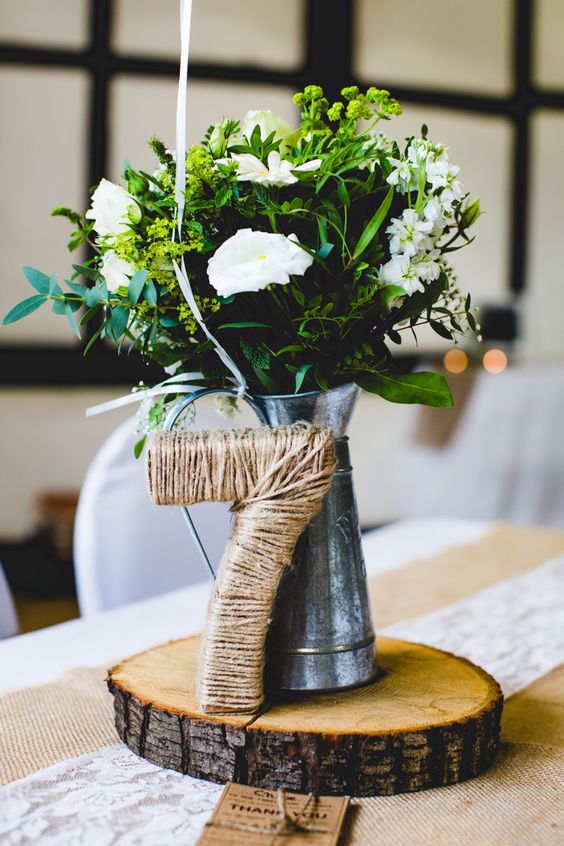 un pichet en étain avec de la verdure et des fleurs blanches, un numéro de table couvert de jute sur une tranche de bois