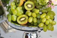 23 a tropical centerpiece with green grapes, kiwi, moss and orchids on a silver stand