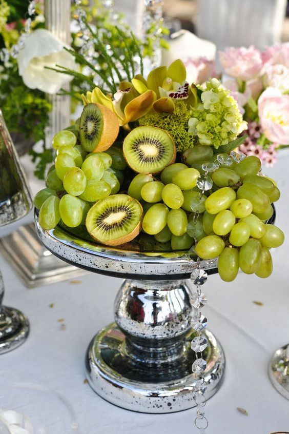 a tropical centerpiece with green grapes, kiwi, moss and orchids on a silver stand