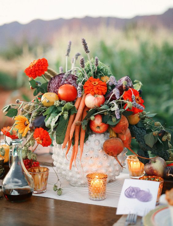 a fall centerpiece with red flowers, apples, carrots, beets, leaves and lavender