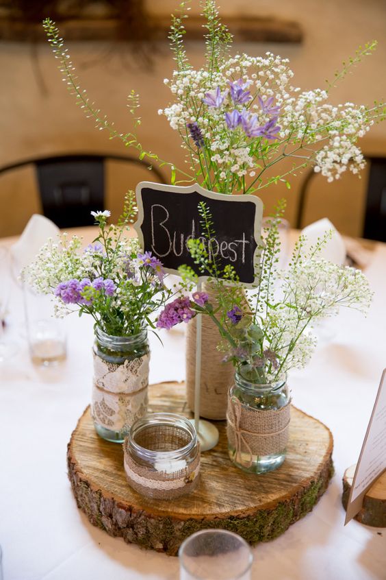a wood slice with jars covered with burlap, jute and lace, wildflowers and a chalkboard table name