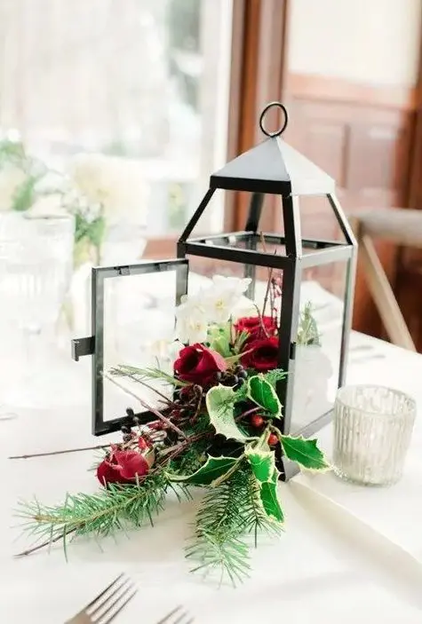a Christmas centerpiece of a lantern and holly leaves and berries, white and red blooms and evergreens