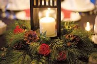 a Christmas centerpiece of a wreath of evergreens, pinecones and red roses and a lantern with a candle
