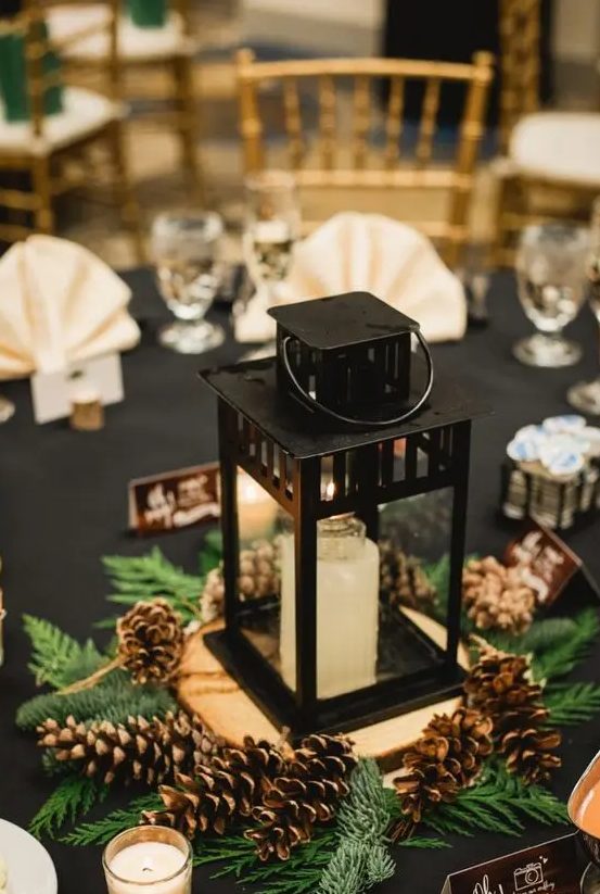 a beautiful winter wedding centerpiece of ferns, pinecones, a candle lantern placed on a wood slice and more candles around is a cozy idea