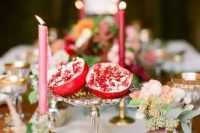 a cluster wedding centerpiece of neutral blooms, greenery, burgundy candles and a cut pomegranate on a silver stand is a gorgeous idea