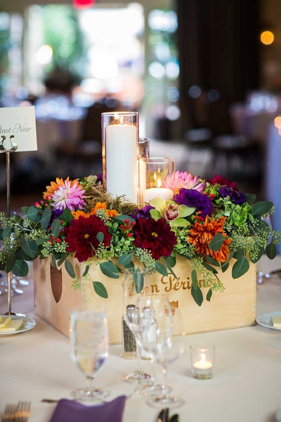 a colorful wedding centerpiece of a crate with super bold blooms and greenery and candles in tall glasses is cool