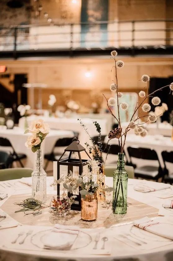 a cool eclectic wedding centerpiece with amber and green bottles, fresh blooms and greenery, a candle lantern is a lovely idea