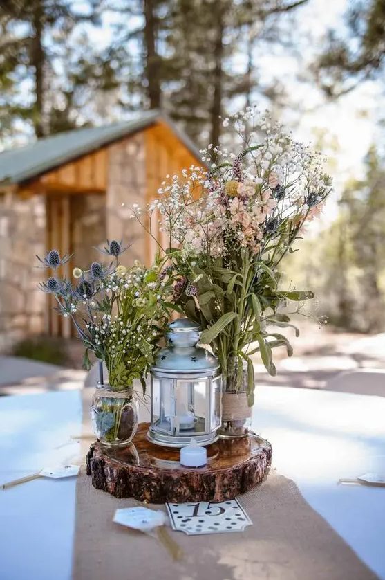 a cozy wedding centerpiece of a wood slice, a hurricane lantern, jars with wildflowers and thistles and a small candle