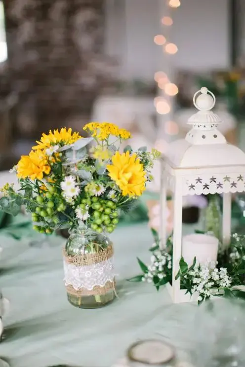 a jar wrapped with burlap and lace and white and yellow blooms, a white candle lantern with greenery and white flowers