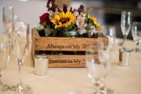 a rustic fall wedding centerpiece of a crate, sunflowers, burgundy and deep purple blooms, greenery and candles around