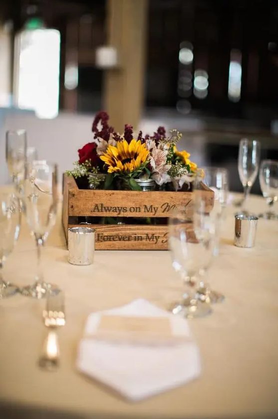 a rustic fall wedding centerpiece of a crate, sunflowers, burgundy and deep purple blooms, greenery and candles around