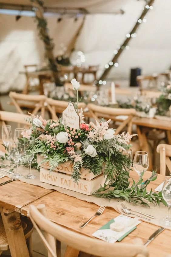 a rustic wedding centerpiece of a crate with greenery, neutral blooms and berries and a table name