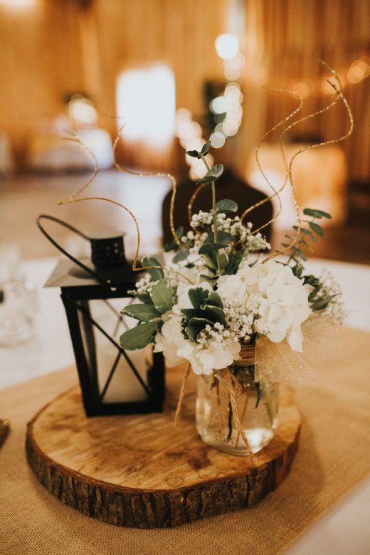 a rustic wedding centerpiece of a tree slice, white blooms with twigs, a lantern is a cozy and simple idea